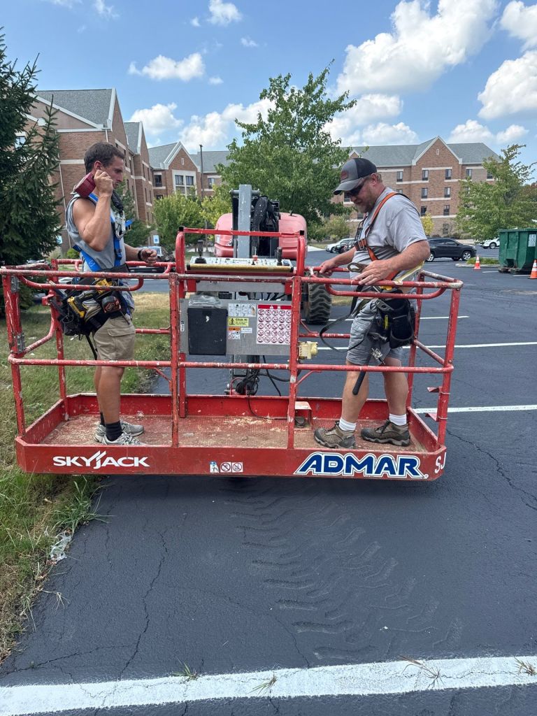 Photograph of two male utility workers in a red SkyJack aerial lift, wearing harnesses, working in a suburban parking lot.